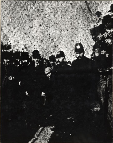 Photograph showing an indistinct image of two policemen with a man in a bowler hat and groups of men either side; the face of a boy can be seen between a policeman and the man in the bowler hat; the photograph has been described as Police Eviction