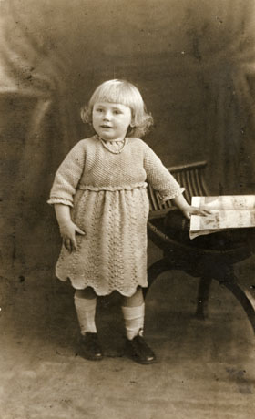 Photograph of a small girl, aged approximately four years, standing with her hand on a publication lying on a piano stool; she is wearing a knitted dress and light socks and dark shoes; the knitting has been identified as Fan and Feather Stitch; the photograph is being taken in a photographer's studio