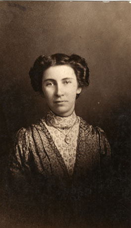 Photograph of the head and shoulders of a young woman wearing a dress of patterned fabric with a long V neck and another patterned fabric inset in the V; she is wearing a locket round her neck; her hair is worn in rolls round her head
