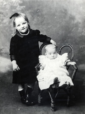 Photograph showing a small girl, aged approximately six years, wearing a dark dress with a high collar and long sleeves; she has a string of pearls round her neck and is wearing shiny boots; she is standing next to an infant sitting in a wicker chair and wearing a light frilly frock