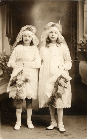 Photograph of two young girls, aged approximately ten and eleven years, wearing light frilly frocks to the knee, light socks to the knee and light shoes; they are also wearing head dresses of net and are carrying bouquets of flowers; behind them are flowers and part of a window; they have been identified as Two Young Bridesmaids