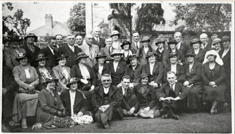 Photograph of approximately thirty eight people dressed in suits, dresses and hats, posed in four rows with a garden wall and trees behind; a woman on the right is wearing a nurse's headdress