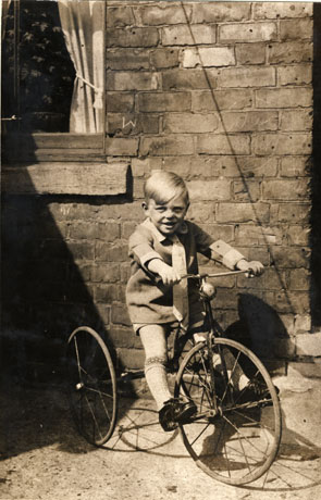 Photograph of a small boy, aged approximately three years, sitting on a tricycle; he is wearing a long shirt, a tie, long socks and shoes; behind him are the wall of a house and part of a window