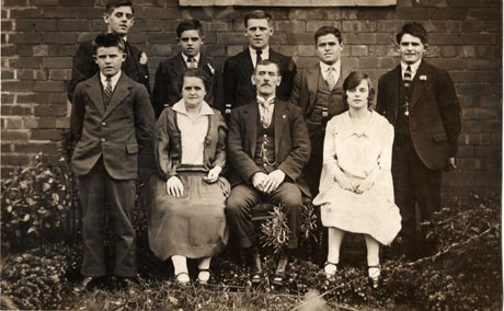 Photograph of a middle-aged man, wearing a suit, tie and wing collar, and a middle-aged woman, wearing a dress with a drop waist, sitting in a garden with five young men behind them and a young man on the left and a young woman on the right; the young men are wearing suits and the young woman a dress