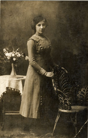 Photograph of a young woman standing with a table, on which there are a cloth and a vase of flowers, behind her; she is resting her hands on the back of a wicker chair; she is wearing a pinafore dress with decorative rows of large buttons on it; underneath that she is wearing a blouse with patterned fabric at the neck and cuffs