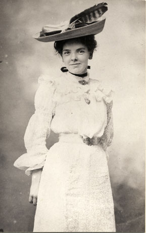 Photograph of a young woman wearing a large hat, with a large feather, on the top of her head; she is also wearing a blouse with a high collar, two frills across the top of the sleeves and across the chest; the sleeves are full and have frills at the wrist; she is wearing a belt with a metal buckle and a straight skirt