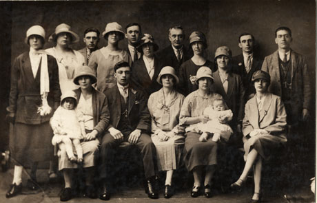 Photograph of ten women and seven men posed in a group; the women are wearing suits, dresses and cloche hats; the men are wearing suits and ties; two women on the front row are holding infants; the group has been identified as a Family Group at a Christening