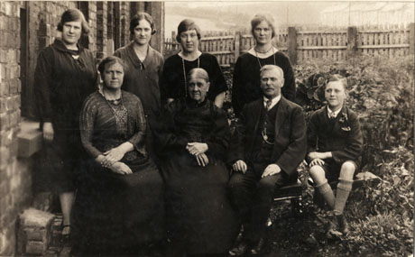 Photograph of a middle-aged man and a middle-aged woman sitting with an elderly woman between them; behind them are four young women, dressed in frocks; on the right is a small boy, aged approximately eleven years, wearing a suit with shorts; they are sitting in a garden with a house on the left and a fence and houses in the distance