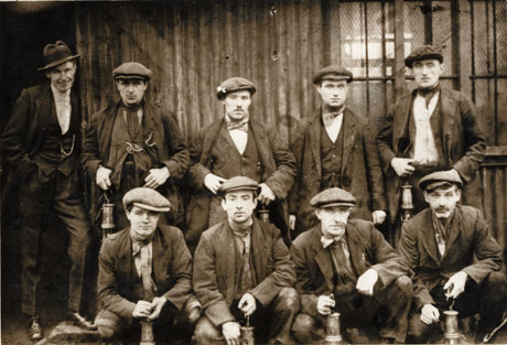 Photograph of eight men, posed in two rows against a corrugated iron building; they are wearing jackets, scarves, coats, waistcoats and are carrying miner's lamps; a man, wearing a suit and a trilby hat, is standing at the left of the picture; they have been identified as miners