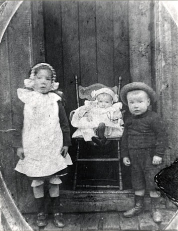 Photograph of a small girl, aged approximately six years, wearing a bonnet, pinafore, dress, socks and shoes; she is standing next to an infant, wearing a frilly dress, in a high chair; on the other side of the infant, is a small boy, aged approximately four years, wearing a straw hat, a jumper and long shorts and boots; they are standing in front of a wooden door