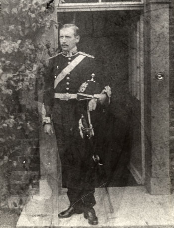 Photograph of a man, standing in front of a door to a house, wearing a military uniform; he has on a tunic with epaulettes, decorations at the wrist, large buttons, a decorative belt and a sash; he is holding a helmet with a badge on the front and a spike on the top; he is holding a sword; he has been identified as William Armstrong
