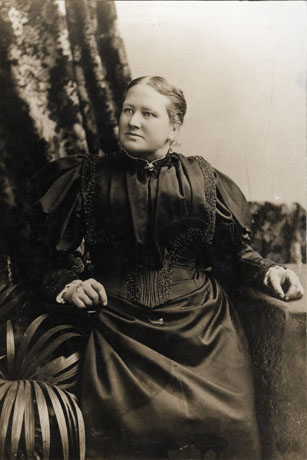 Photograph of a woman wearing a dark dress with puffed sleeves and tucks and embroidery on the bodice; her skirt is long, plain and straight