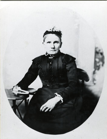 Photograph of a woman sitting with her right hand holding a book and resting on a table; she is wearing a dark cap, a dark bodice with tucks and straight sleeves; she is wearing a brooch at her throat; she has a dark full skirt