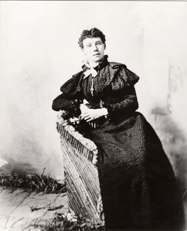 Photograph of a woman sitting on a bench wearing a dress with mutton sleeves and frills over the shoulder; there is a bow at her neck; the dress has a dark lace over a dark under dress