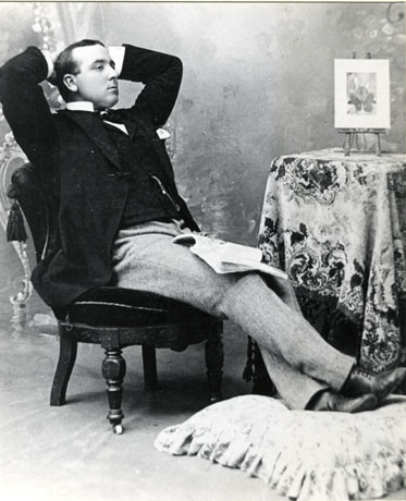 Photograph of a young man sitting at his length on a padded chair with his arms behind his head; a table, covered in a patterned cloth and bearing a miniature easel on which there is a picture, is on the right of the photograph; the young man's feet are resting on a cushion; he is wearing a dark jacket and waistcoat with a wing collar and a watch chain; he has light narrow trousers and dark boots