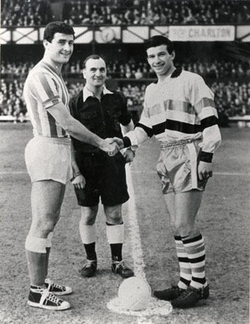 Photograph showing a referee, dressed in black shirt and shorts, standing between two footballers in football strip shaking hands over a football in a stadium; stands full of spectators can be seen behind the three figures, one of whom has been identified as Stan Anderson