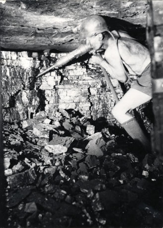 Photograph of a man stripped to the waist wearing shorts, socks and a helmet, working at a coal face with an automatic tool, which has been identified as a pick; the roof and wall of coal can be see in detail