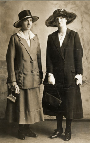 Photograph of a young woman wearing a dark hat with a high crown, a jacket with a pattern of large buttons on the lapels and on the pockets at the bottom on either side of the front; she is also wearing a full skirt and shoes; she is carrying a small handbag and a pair of gloves;she is accompanied by another young woman who is wearing a hat with a lower crown and a wider brim; she is wearing a long plain dark jacket, a full dark skirt, dark stockings and dark laced shoes with a heel; she is also wearing light-coloured gloves and is carrying a large handbag