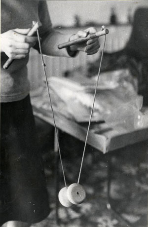 Photograph showing a pair of hands holding sticks which are joined by a string on which a bobbin is being reeled; behind the bobbin, part of a person in a skirt and jumper, a carpet and a chair, can be seen; the bobbin and sticks are described as Diabolo