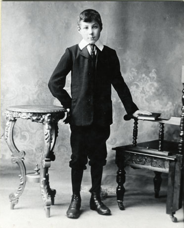 Photograph of a boy, aged approximately ten years, standing with his right hand on a small occasional table and his left hand on the arm of a chair in a photographer's studio; he is wearing an Eton collar, a tie, a long dark jacket, knickerbockers, long dark socks, and boots