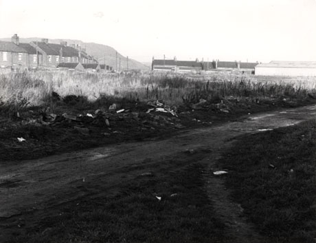 Photograph showing an earth road through scrub, with the rear of a terrace of houses on the left and the side of a hill beyond the terrace; in the centre and right of the picture in the distance, are the rear of another terrace and single-storey, possibly industrial, buildings