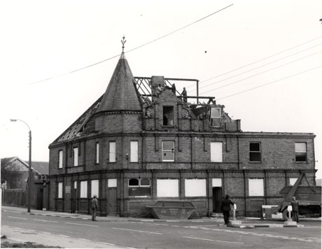 Photograph showing the facade of two sides of a brick building, on the corner of a road, under demolition; the roof has only its laths remaining, apart from a turret at the corner; two men are on the roof and three men are on the ground; two skips with the name, Mercers, of Seaham, are in the road near the building, which appears to have been a public house