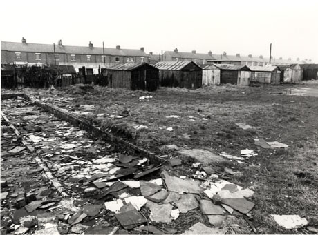 Photograph showing, in the foreground, the shape of the foundations of a building, possibly a garage, with pieces of roofing felt lying on and around the foundations; in the distance, beyond rough grass, are eight sheds, possibly garages, with two terraces of houses beyond them in the distance