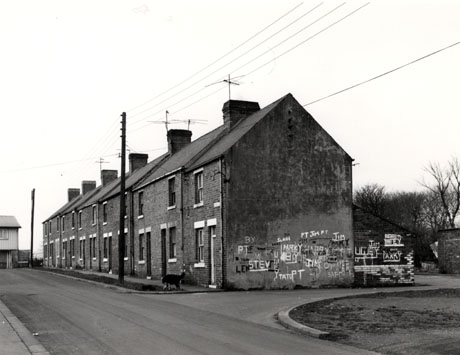 Photograph showing the end and front of a terrace of twelve houses receding from the camera; on the end of the terrace is graffiti, consisting of people's names; the graffiti on the wall of the yard of the end house is the same as that in shot0207