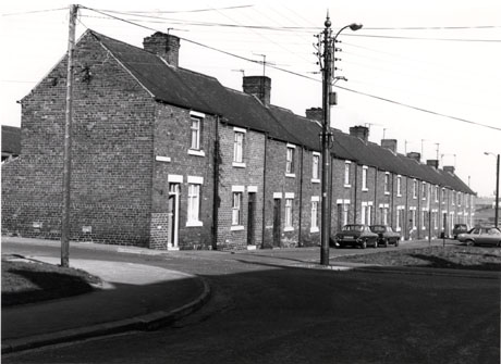 Photograph of the facade of sixteen terraced houses running away from the camera; there are three cars and a van parked in front of the houses