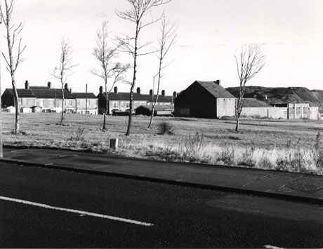 Photograph showing a road running across the picture, in the foreground; beyond that is an open grassed space and, beyond that, there are single-storey buildings with a hill, possibly a pit heap, beyond them; on the left of the picture, in the distance, are two terraces of houses with sheds in front of them