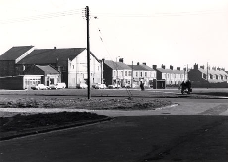 Photograph showing, in the foreground, pavement and grass verges; a small shop, a large building, used as a club, a row of shops at the end of a terrace, can be seen beyond the pavement and verges; the rear of another terrace facing the first terrace can be seen