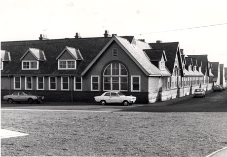 Photograph of the exterior of a large single-storey building with large windows and steep roof with dormer windows in it; five cars are parked in front of the building; it is possible that the building is a school