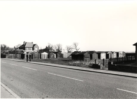 Photograph showing the surface of a road running across the front of the picture; on the far side of the road, fifteen garages for individual cars can be seen, in three rows at right angles to the road; beyond the garages, two large houses can be seen; two men are standing talking on the far side of the road