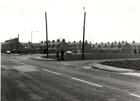 Photograph showing the surface of a road running away from the camera to the right towards the ends of five terraces which abut the road; two cars can be seen on the road and two people can be seen talking on the pavement