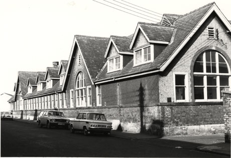 Photograph showing, close-up, the exterior of a large single-storey building with large windows and a steep roof with dormer windows; three cars are parked outside the building, which is, possibly, a school and is the same building as in misc0072