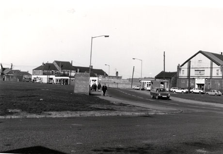 Photograph showing a road running across the front of the picture and turning to the left; at the end of the road are buildings and shops; on the right are two shops and a large building used as a club, in front of which there are six cars parked; a car is driving down the road towards the camera and four people are walking down the pavement towards the camera; the club and adjacent shops are those in misc0069 and misc0079