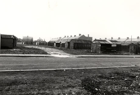 Photograph showing a road running across the photograph on the foreground; beyond the road are huts, possibly garages, in two rows at right angles to the road; beyond the garages the roofs of houses can be seen