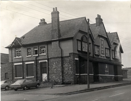Photograph showing the exterior of the front and side of a large brick building with three gables and with boarded-up windows on the ground floor and broken windows on the first floor; the front of the building bears a sign reading: The Albert Inn; two cars are parked at the side of the building