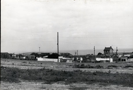 Photograph showing, in the foreground, open rough ground, with fences and sheds in the distance across the road which runs across the middle of the photograph; a coal waggon can be seen in the middle of the photograph beyond the sheds; a house can be seen on the right beyond the sheds