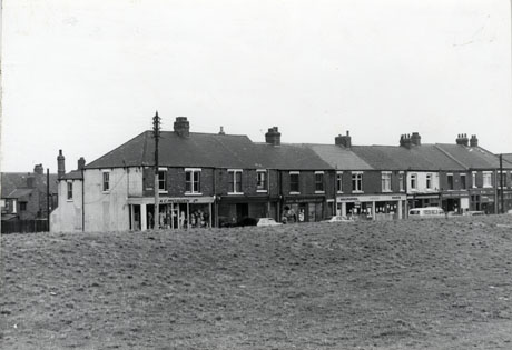 Photograph showing a terrace of shops seen from across a bank of grass in the foreground; the shop on the left is A. C. McCullough Ltd., which appears to be a drapery shop; the next cannot be identified; the next is K. and C. Wilson; the next sells wallpaper and paint, but its name cannot be read as cannot those of the further four which can be seen