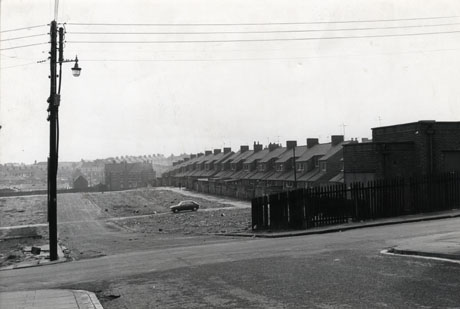 Photograph showing, from a slightly high point, four areas of cleared ground; a road is running across the picture at the front and a road is running off that away from the camera between four squares of cleared ground, each of which would, most likely, have had two terraces separated by a back road down the middle of the plot; on the right of the picture is a surviving terrace; further terraces and other buildings can be seen in the distance