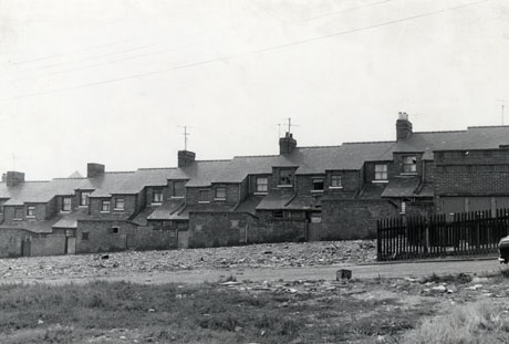 Photograph showing the rear of a terrace of houses across an area of open rough ground, possibly an area where houses have been demolished