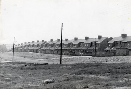 Photograph showing the rear of a long terrace of houses, on the right of the picture, running away from the camera; in the rest of the photograph is cleared ground