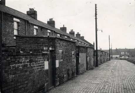 Photograph showing the walls of the yards and backs of outhouses of a terrace of houses; a cobbled road is running in front of the buildings