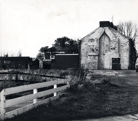 Photograph showing, on the right, the end wall of a house; a wall is attached to the left of the house and another house can be seen beyond it; other buildings are on the left of the picture in the distance; in the foreground of the picture on the left is a low fence surrounding some plants