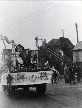 Photograph showing the rear of a lorry dressed with pictures of dragons and daffodils; leeks can be seen at either corner of the back of the lorry; on the lorry are a replica harp and spinning wheel; two women in traditional Welsh costume are standing behind the harp; behind the lorry are four people, and a man in uniform, standing near a wooden footbridge over a railway line, a railway signal and the corner of a railway building