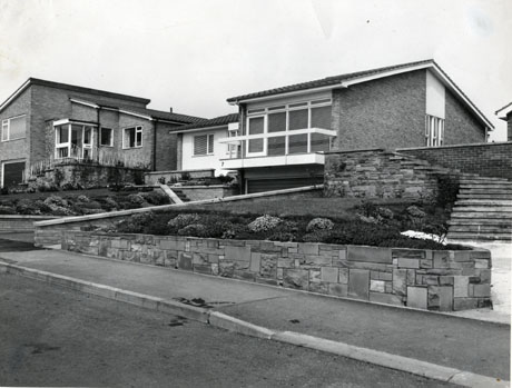 Photograph of the exterior of two Post-Second-World-War bungalows taken from the road outside; they have large windows, gardens with Alpine plants, tiles on the roofs, steps and metal fences