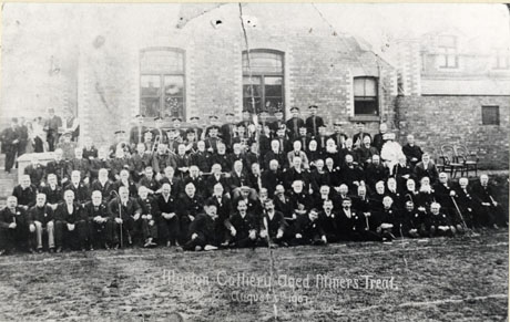 Photograph of a group of seventy two elderly men posed in front of a large brick building, with twenty three bandsmen and a clergyman behind them; six people can be seen in the distance standing at the side of the building; the photograph has the following words printed on it: Murton Colliery Aged Miners Treat, August 1907