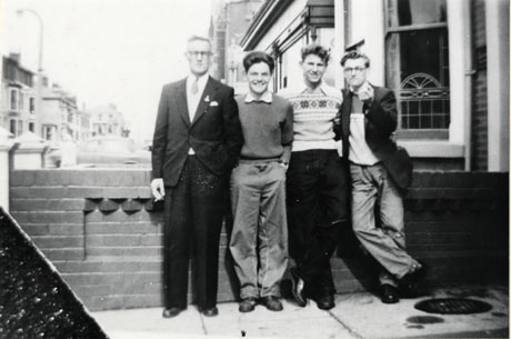 Photograph of a middle-aged man and three young men standing in front of a small wall dividing the front gardens of houses; behind the men, the bay window of the next house can be seen, and large houses, possibly hotels, can be seen in the distance further along the street; the photograph has been identified as Bobby Burns at Blackpool