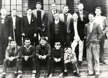 Photograph of fourteen men and two boys sitting on the steps of a brick building; one of the boys is holding a cricket ball and all are wearing ordinary clothing; the photograph has been identified as Murton Democratic Cricket Club 1965